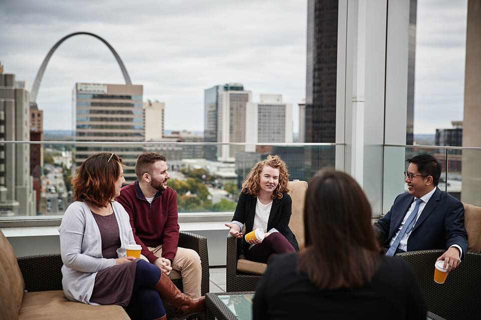 A group of students and staff sit on the roof top of the SLU Law school talking around a table with the St. Louis skyline and Arch in the background. 