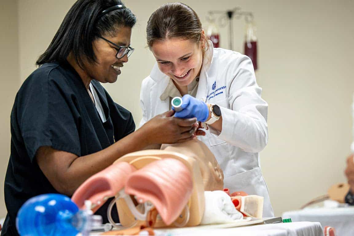 A SLU student practices intubating a sim mannequin while an instructor looks on.