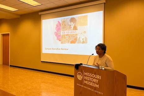 A speaker stands at a lectern that reads Missouri History Museum, with a projection screen in the background.