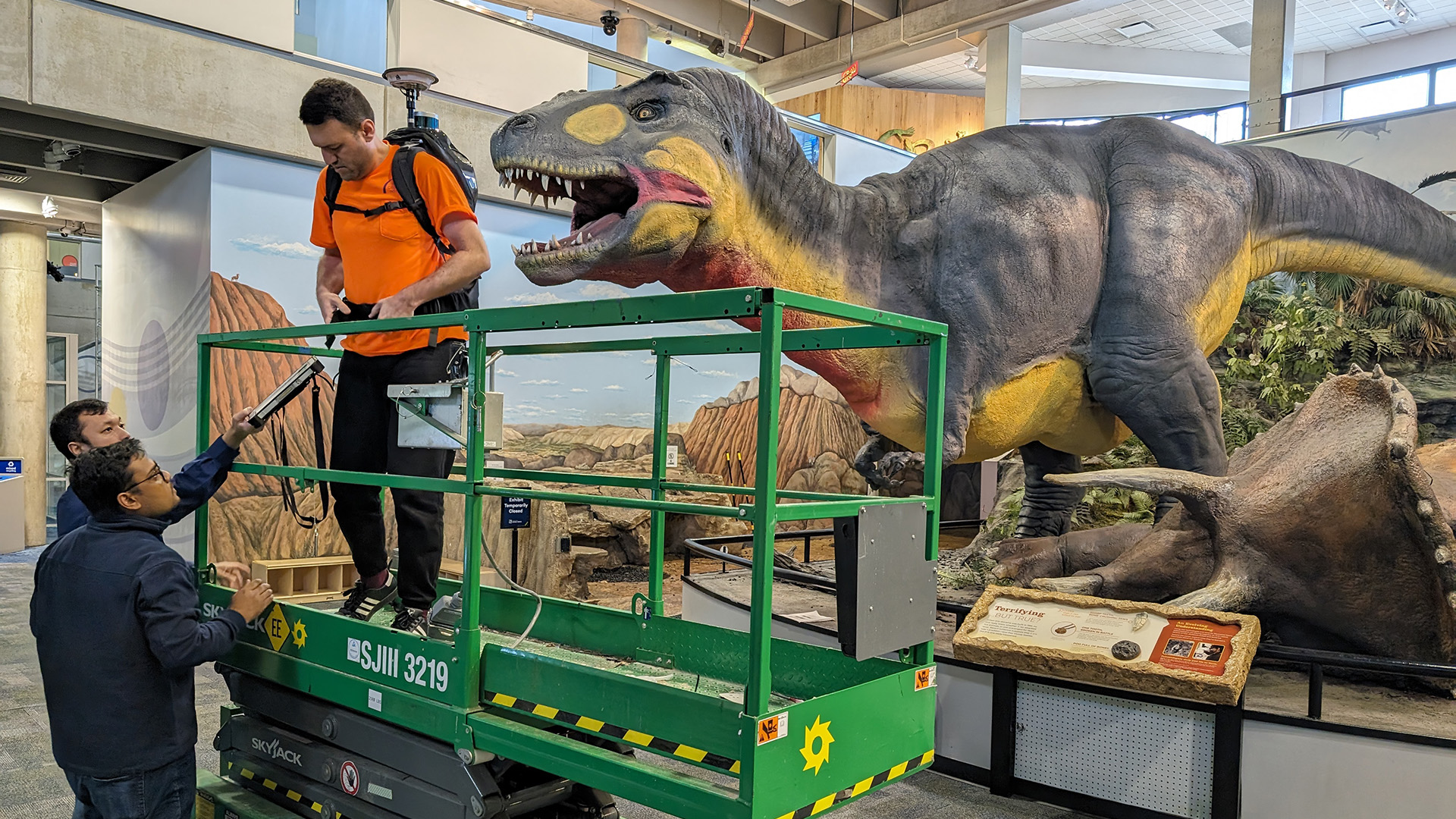 Cagri Gul, master's student in geographic information science, gets situated with the LiDAR backpack scanner before beginning to scan the repaired T. rex. Also pictured: Alifu Haireti, geospatial computing engineer with TGI, and Mustafizur Rahaman, master's student in geographic information science.