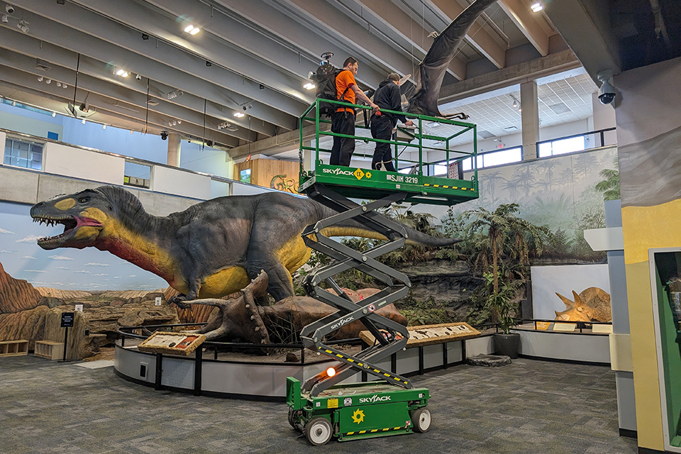 Cagri Gul, master’s student in geographic information science, stands atop the SkyJack as Chris Lucas, exhibit designer with the Saint Louis Science Center, drives. The scanning team needs to capture angles of the diorama from above in order to create a more complete scan; simply walking with the backpack on the ground would not collect data from the top of the dinosaurs.