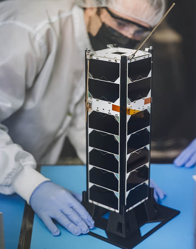 A small satellite with a height of about about 30 centimeters long and 10 centimeters on its side shaped like a metal loaf of bread sits on a table. A student wearing protective gear is seen in the background.