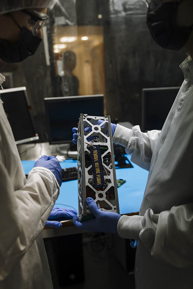 Undergrad engineering students working on engineering precursor, “Copper 2." The hands of a student are seen holding a small satellite that has a tag with the words "remove before flight" on it.
