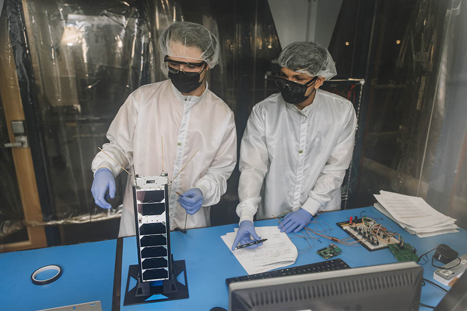 Two students wearing protective coats, face masks, gloves and head wear, examine a piece of equipment on a table.