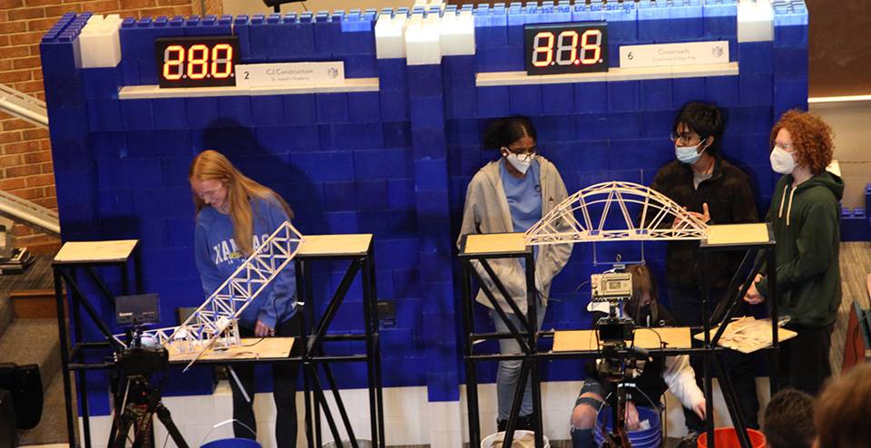two student teams watch as one balsa wood bridge collapses under the weight