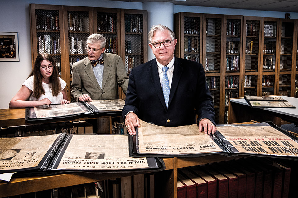 A female student and male professor examine historic newspapers in the background, while SLU alumnus Tim Drone stands with another newspaper in the foreground.
