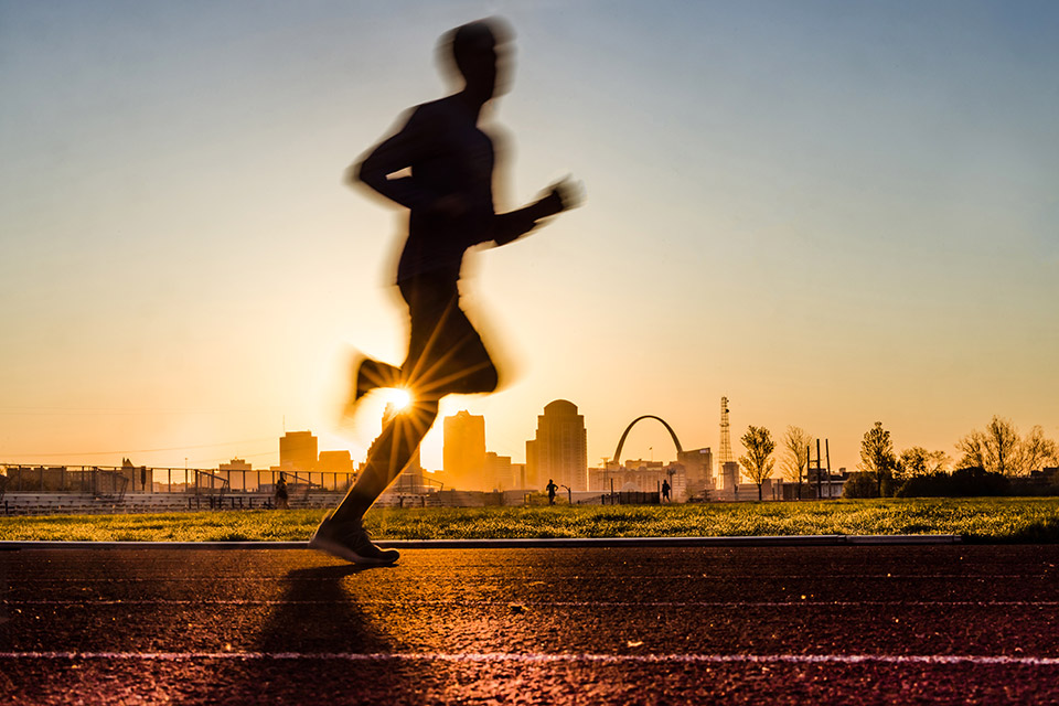 A silhouette of a runner at dawn on the track at SLU's Medical Center Stadium, with the Gateway Arch and the St. Louis skyline in the background