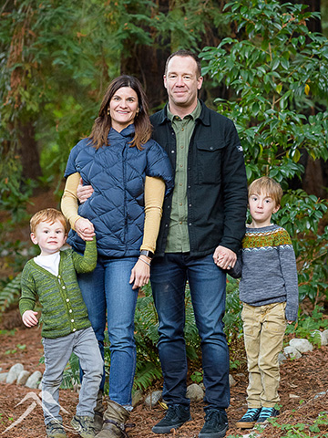 Tim Bantle's family portrait. Two young boys in sweaters flank Sara, who wears a navy quilted jacket and jeans, and Tim, who wears a green shirt, black jacket, and dark jeans. They stand in a forest setting.
