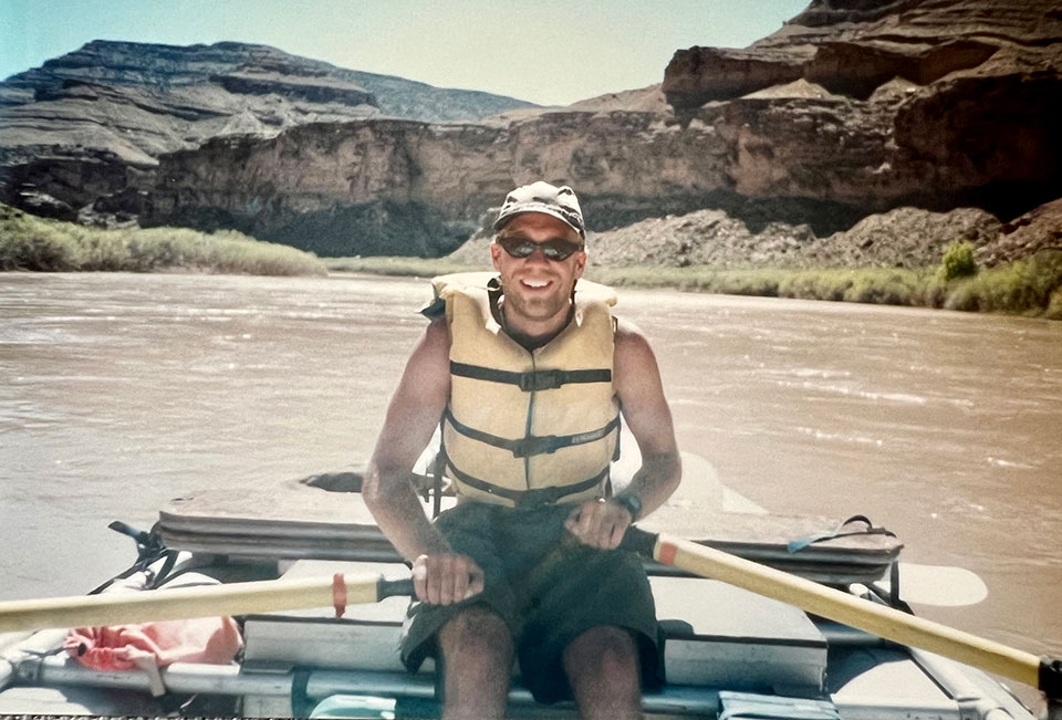 A man in his 20s paddles an oar rig on a brown river with the Rocky Mountains in the background.