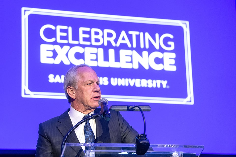 Dr. Richard A. Chaifetz speaks during a gala standing in front of a projection reading Celebrating Excellence, Saint Louis University