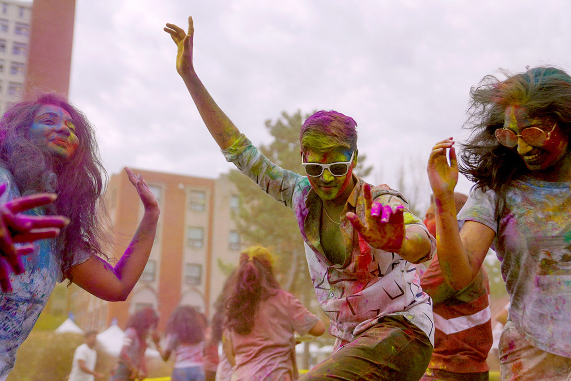 Three college students celebrate Holi by dancing on the quad on SLU's campus. They are covered in colorful powders. A male student wearing white sunglasses raises his arms, dancing. He is flanked by two dancing females.