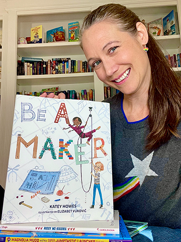Author Katey Howes, a middle-aged woman with long dark hair, wears a sweater with a star on it and holds her children's picture book, Be a Maker.