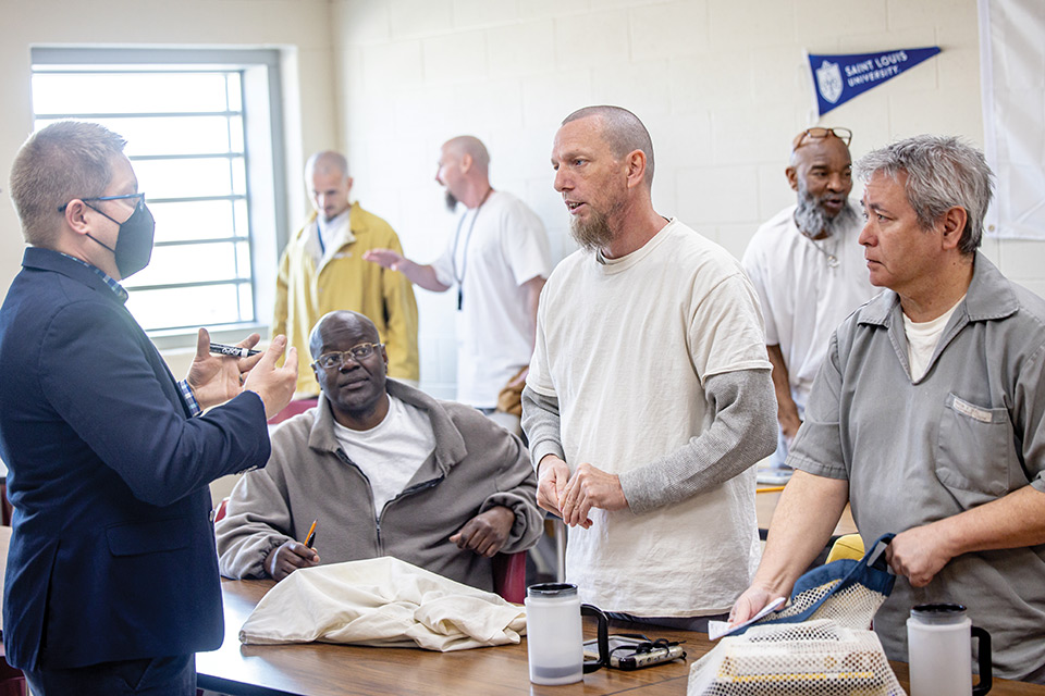 Three incarcerated adult male students talk with their professor. The professor (left) is a white man with short blondish hair. He wears a navy sport coat and a black mask over his mouth. Several other incarcerated classmates talk in the background.