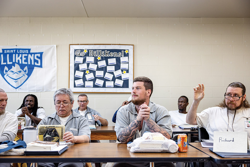 Incarcerated male students sit at long tables during class. An older man with gray hair and glasses looks at his tablet. A younger man with tattooed forearms looks to the professor. A man with long brown hair and a long beard raises his hand. 
