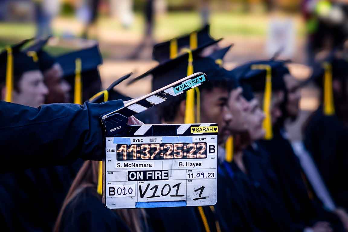 SLU Goes Hollywood A clapperboard with the words "ON FIRE" is positioned in front of a crowd of actors wearing graduation caps and gowns.