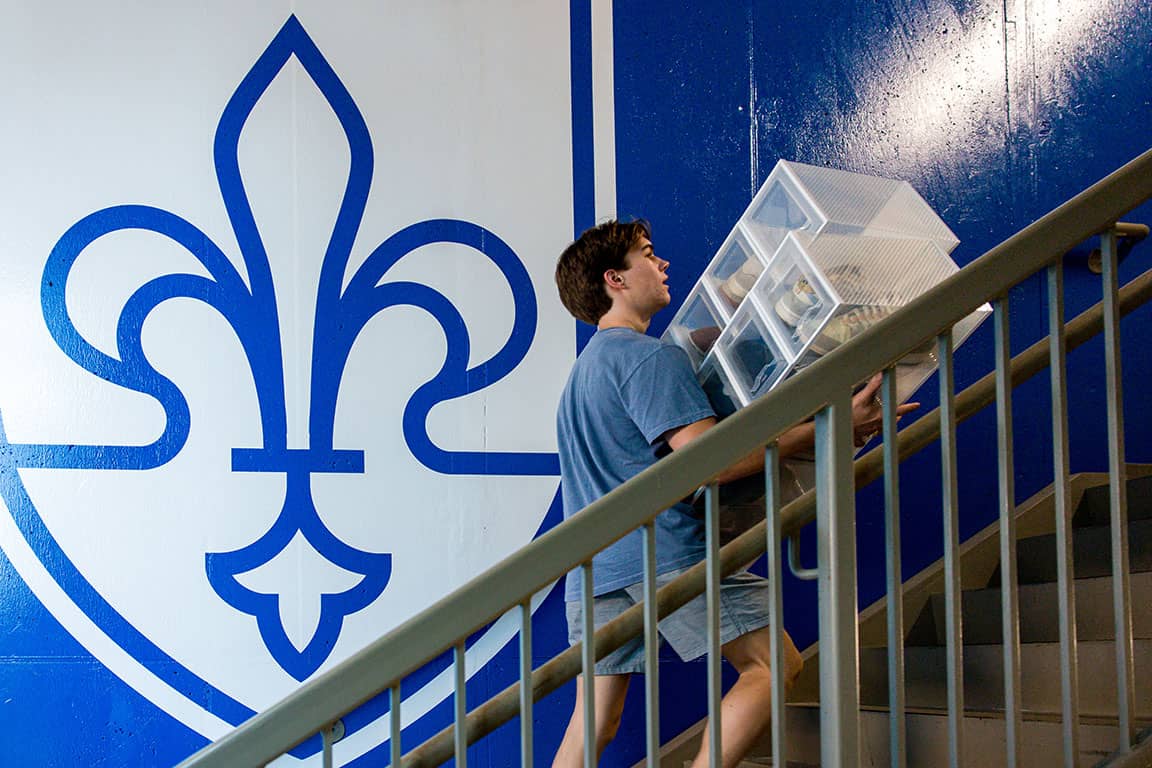 A student carries their belongings up the stairs in front of a mural of the SLU shield during move-in
