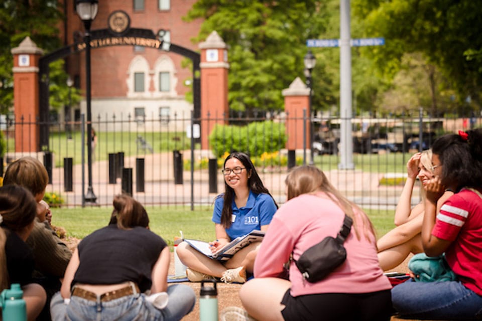 A SLU 101 leader chats with admitted students while sitting in a circle on a SLU lawn