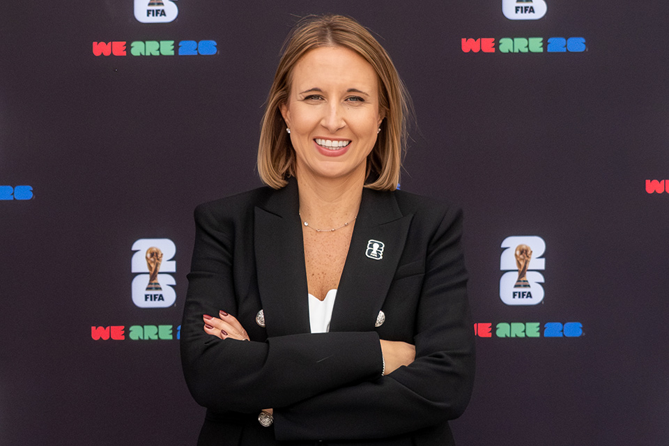 A young white woman with shoulder-length straight blonde hair wears a black pantsuit and crosses her arms while standing in front of a backdrop marked with the FIFA logo.