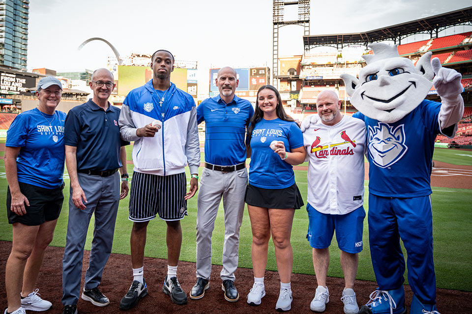 A group of people stand with the Billiken mascot on the field at Busch Stadium