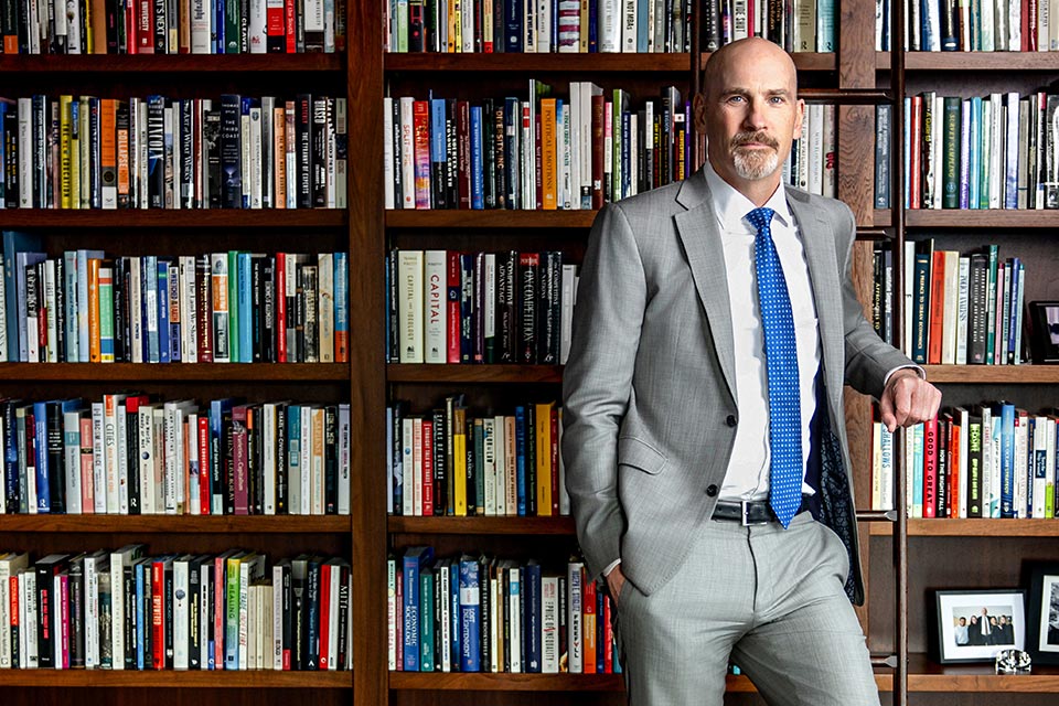 A bald white man in a grey suit stands in front of a wall-length bookcase full of books.