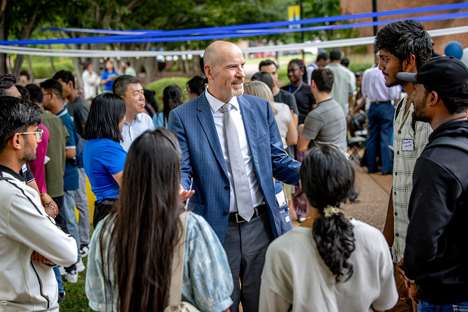 A tall, bald white man in a blue suit is encircled by several college students