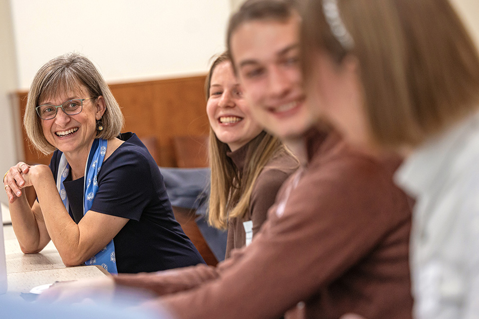 A white woman with short grey hair with bangs and glasses smiles and sits at a table with several students.